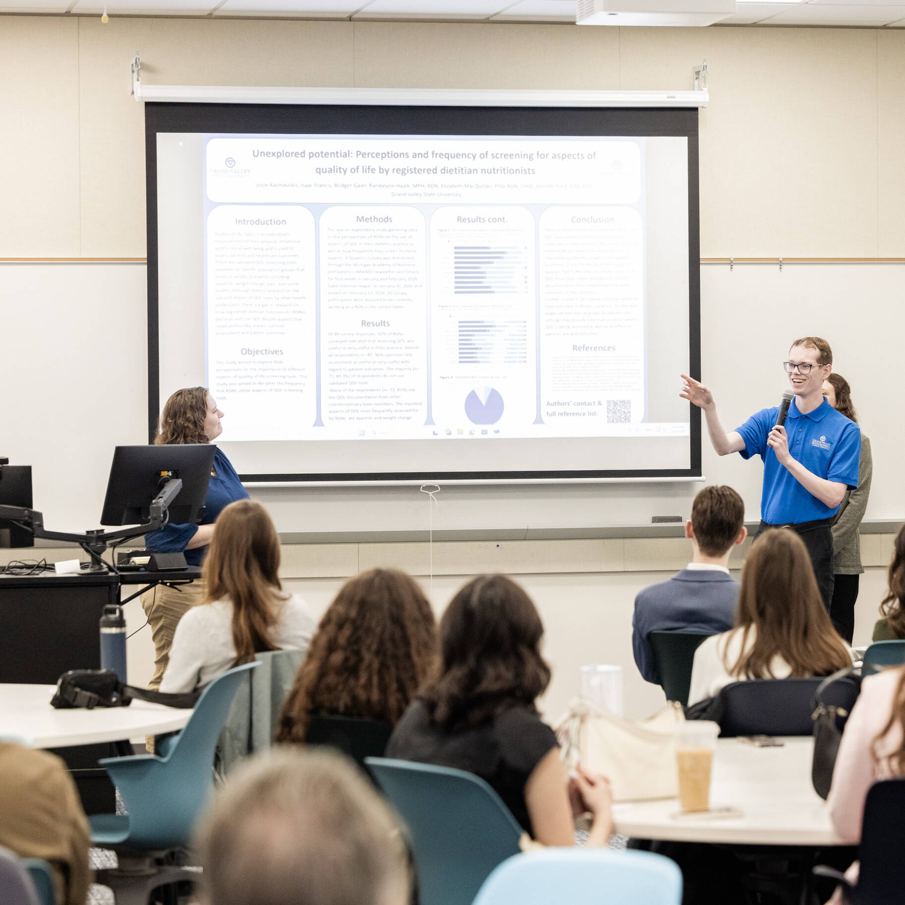 A smiling student gestures to a poster on a large screen, as his group presents their capstone project during Clinical Dietetics Research Day.
